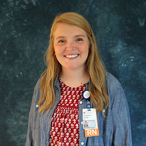 Smiling woman with long blonde hair wearing a red patterned top, blue jacket, and ID badge, standing in front of a dark blue backdrop.