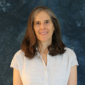 A headshot of a smiling woman with long brown hair, wearing a patterned short-sleeve blouse against a mottled blue backdrop.