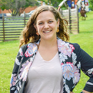 A smiling woman with curly brown hair, wearing a colorful floral blazer, stands outdoors in a grassy area with a wooden fence in the background.