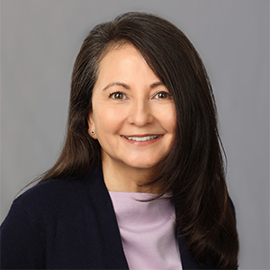 A smiling woman with long, dark hair wears a navy cardigan and a light purple top against a gray background.