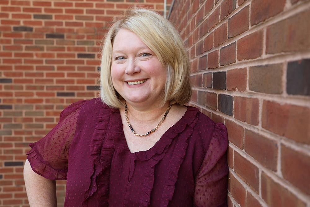 Louise Fleming head and upper torso. She is smiling warmly, is wearing a burgundy blouse, has shoulder length blond hair, and is standing in front of a brick wall.  She is smiling.