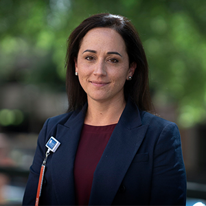 a woman with dark brown hair and blue blazer smiling at camera with blurred background