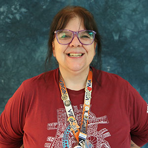 A woman smiling with glasses wearing a red graphic tee shirt against a blue mottled background.