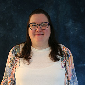 A woman smiling with glasses wearing a white shirt with a floral cardigan against a dark blue mottled background.