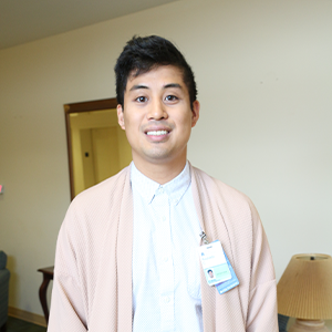 A young man with dark hair smiles for a photo indoors. He is wearing a light pink cardigan over a collared shirt and has an ID badge clipped to his cardigan.
