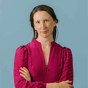 Professional headshot of a confident woman with long brown hair, wearing a vibrant magenta and black patterned blouse, standing with her arms crossed against a solid light blue background.